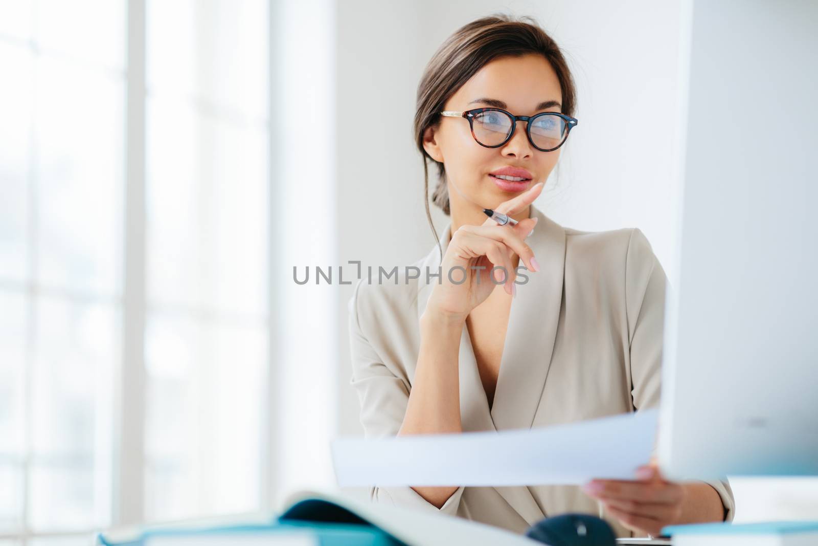 Female economist poses in coworking space, focused in monitor, verifies or checks figures from financial report, holds paper document and pen, has dark combed hair, wears formal business suit by vkstock