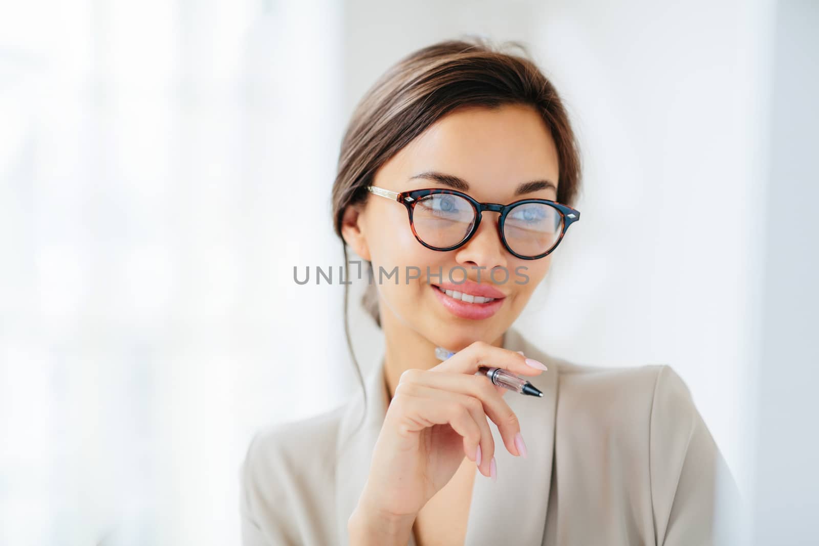 Close up shot of successful tender young woman with combed hair, keeps hand near chin, holds pen, focused in monitor, prepares business report, has manicure, dressed elegantly, works indoor.