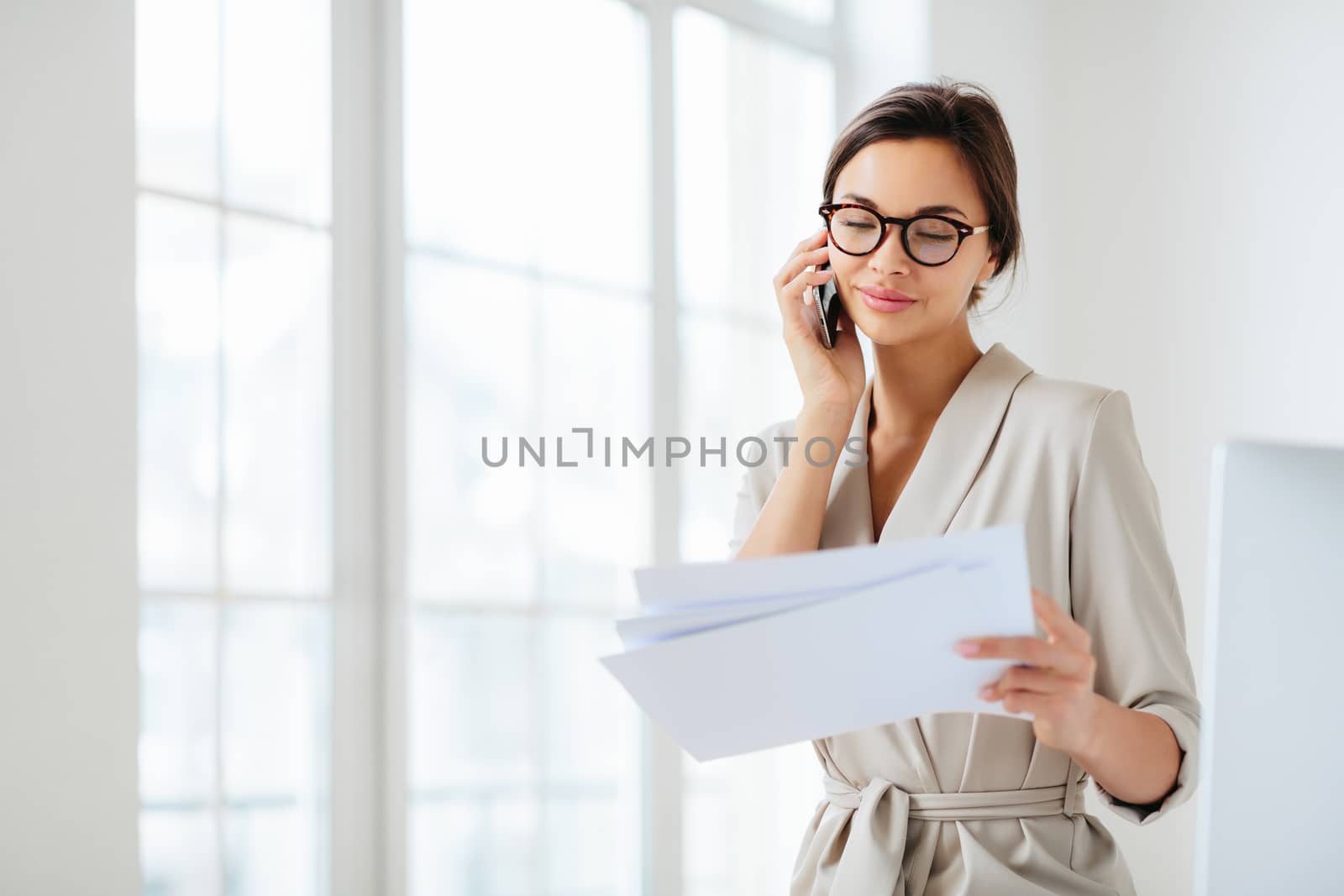 Horizontal shot of business lady discusses details of contract, works in office, concentrated on information from accountings, holds paper documents during telephone conversation, dressed formally by vkstock