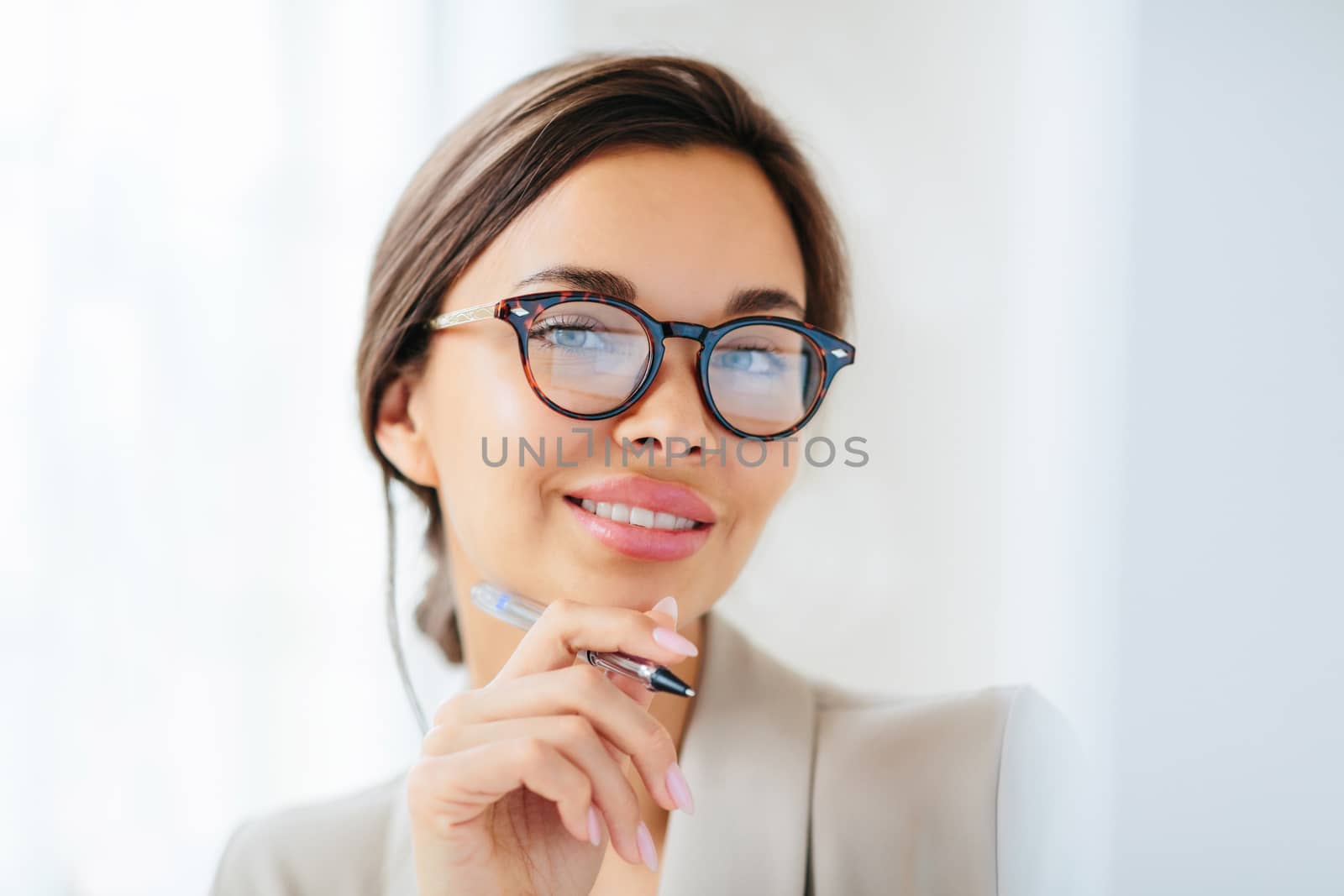 Pleasant looking young female model with makeup and manicure holds pen, touches chin gently, looks directly into camera, dressed in formal clothing, works in office of company, wears optical glasses by vkstock