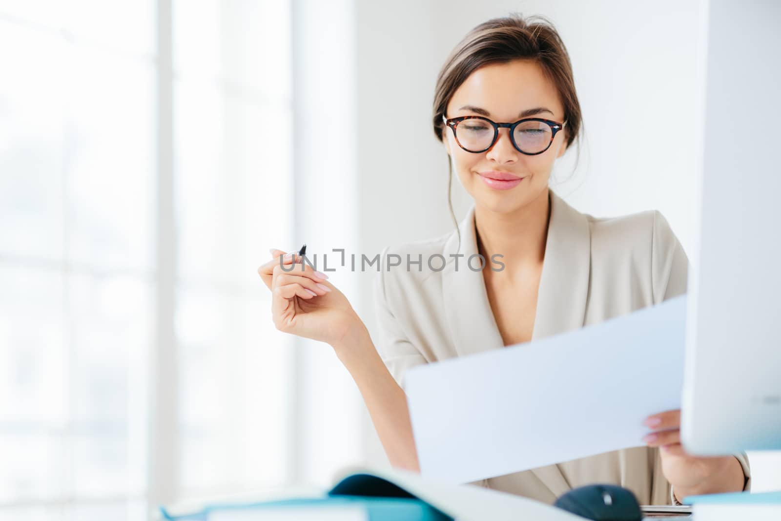 Concentrated successful businesswoman looks attentively in paper, studies terms of contract, holds pen, writes in documentations, dressed formally, poses at desktop against white spacious interior