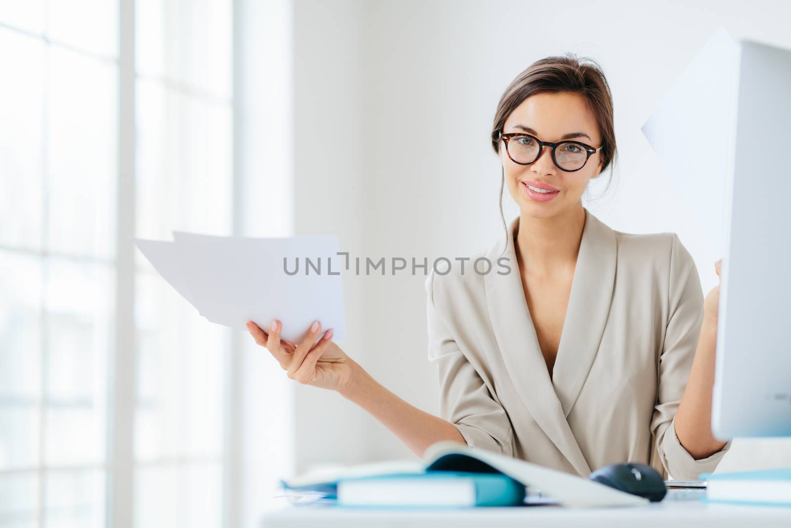 Indoor shot of good looking female in formal suit, holds paper documents, works on organization and creating startup project, poses at desktop with computer, checks information for presentation