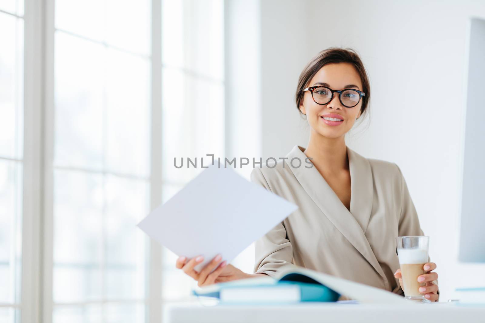 Photo of delighted dark haired European woman in transparent glasses, works with documents, reads information data, dressed in elegant clothing, drinks milkshake sits at desktop in office makes report