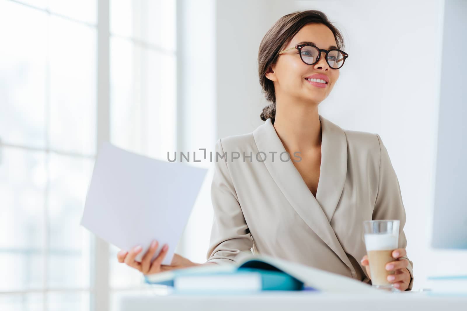 Cheerful businesswoman reads paper documents, focused aside with smile drinks milkshake sits at desktop makes business plan for project. Female executive manager in spectacles poses in coworking space by vkstock