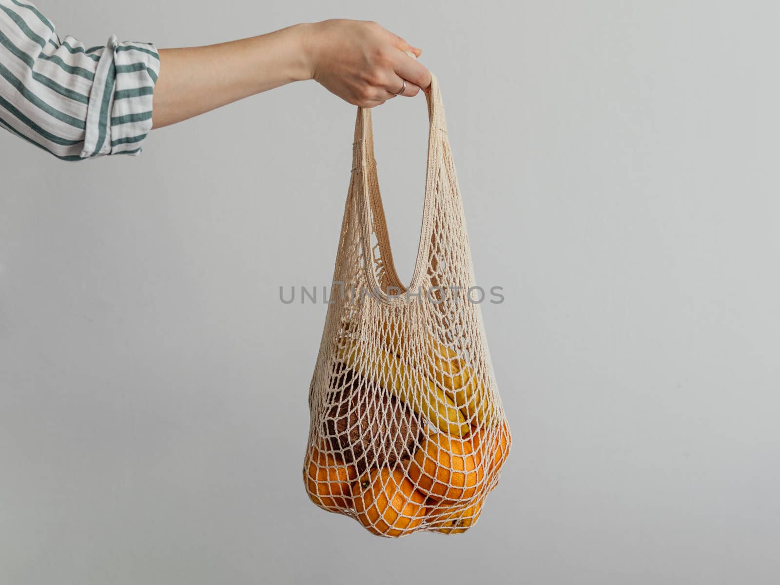 Mesh bag with fruits in female hand. Stylish young woman hand hold mesh shopping bag on light gray wall. Modern reusable shopping, zero waste concept.