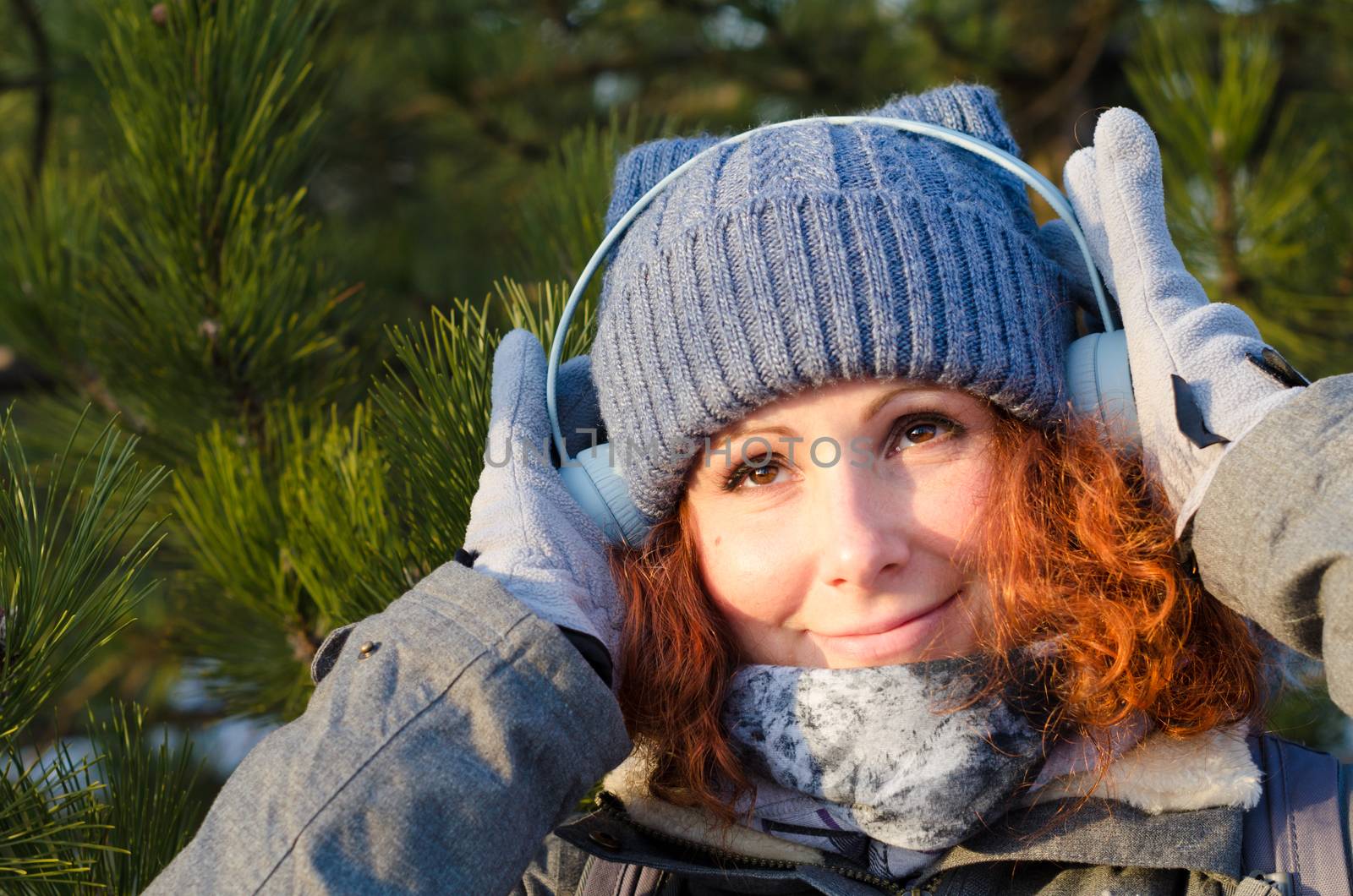 red-haired young girl in winter clothes listens to music with headphones on the background of pines
