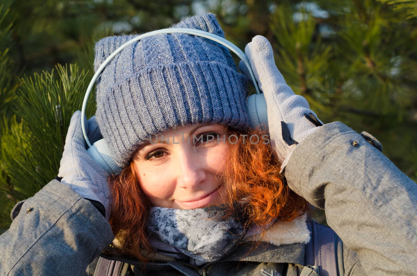 red-haired young girl in winter clothes listens to music with headphones on the background of pines