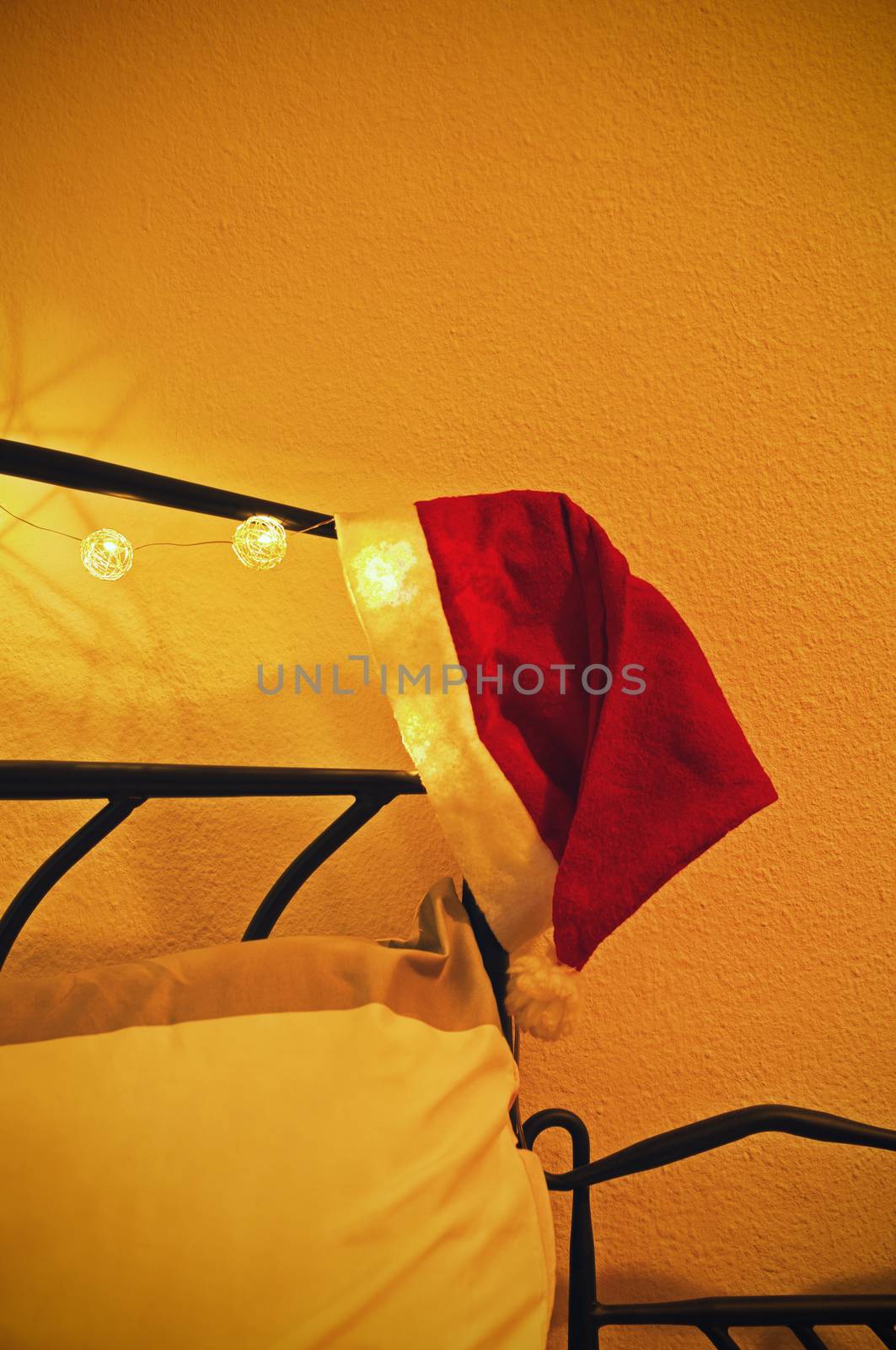 Christmas eve concept. Santa’s hat placed at the edge of a wrought iron bed frame, decorated with garland of Christmas lights. Coziness, comfort, interior and holidays concept.