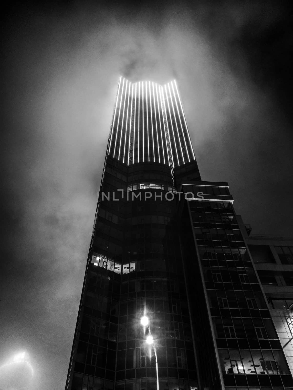 A black and white photograph of a skyline building illuminated by lights, covered in misty pollution fog. Bottom up or ground up view of a tall building covered in thick fog late at night.