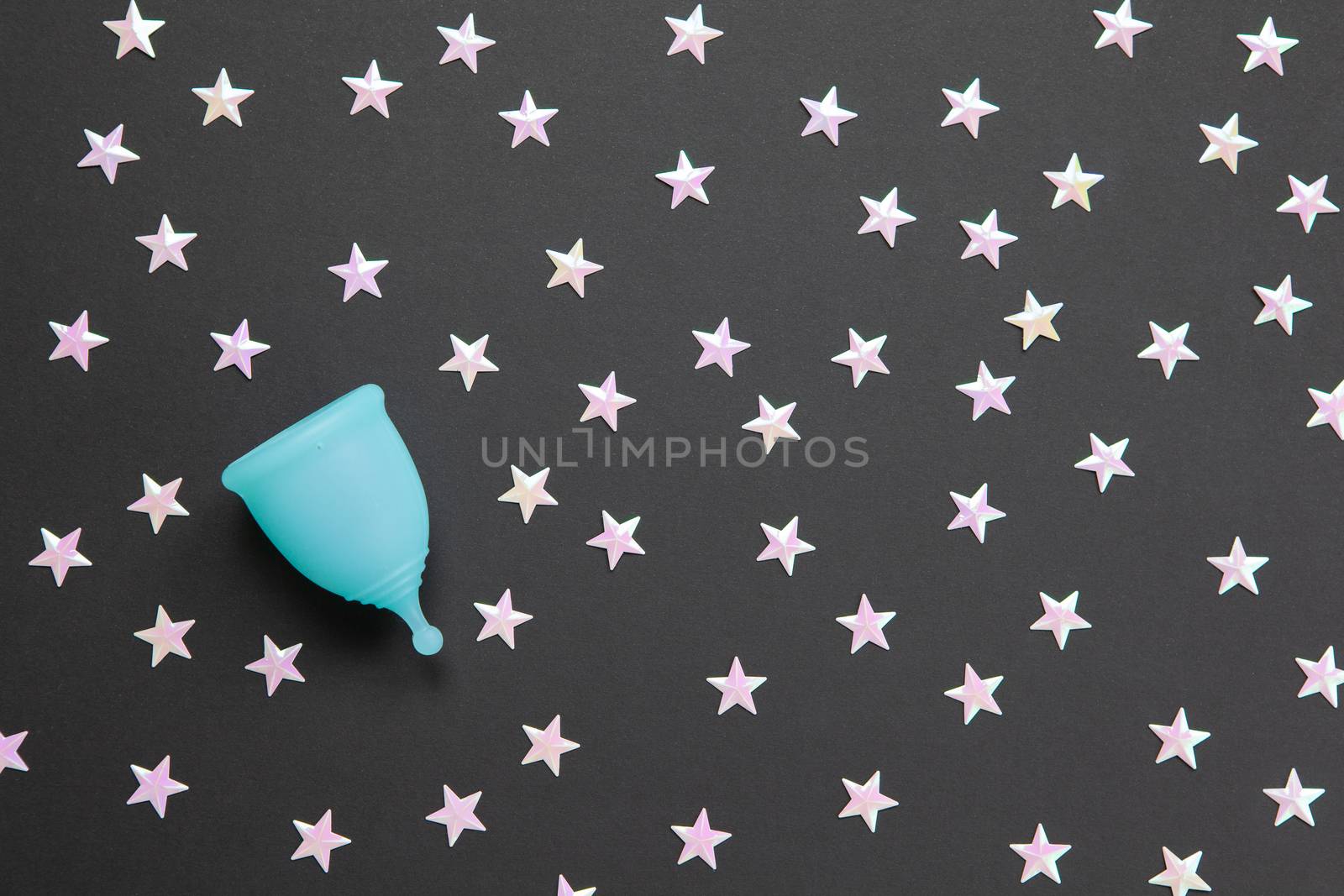 Turquoise menstrual cup on black background with numerous stars. Concept comfort of using cup at night, zero waste, savings, minimalism. Feminine hygiene product, flat lay, copy space. Horizontal.