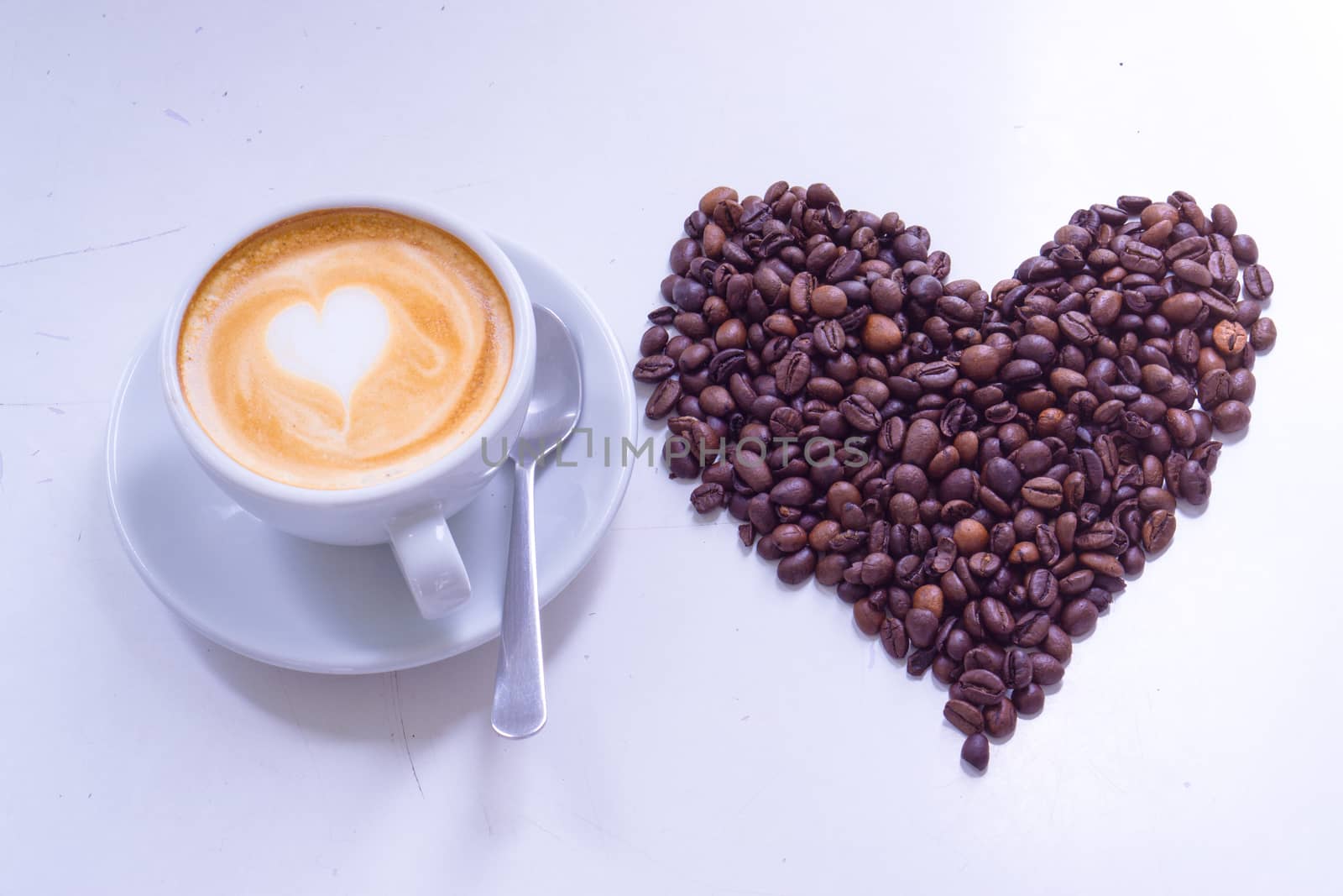 White cup of coffee latte with white heart shape milk foam and heart pattern coffee beans on white table. top view, copy space for text.