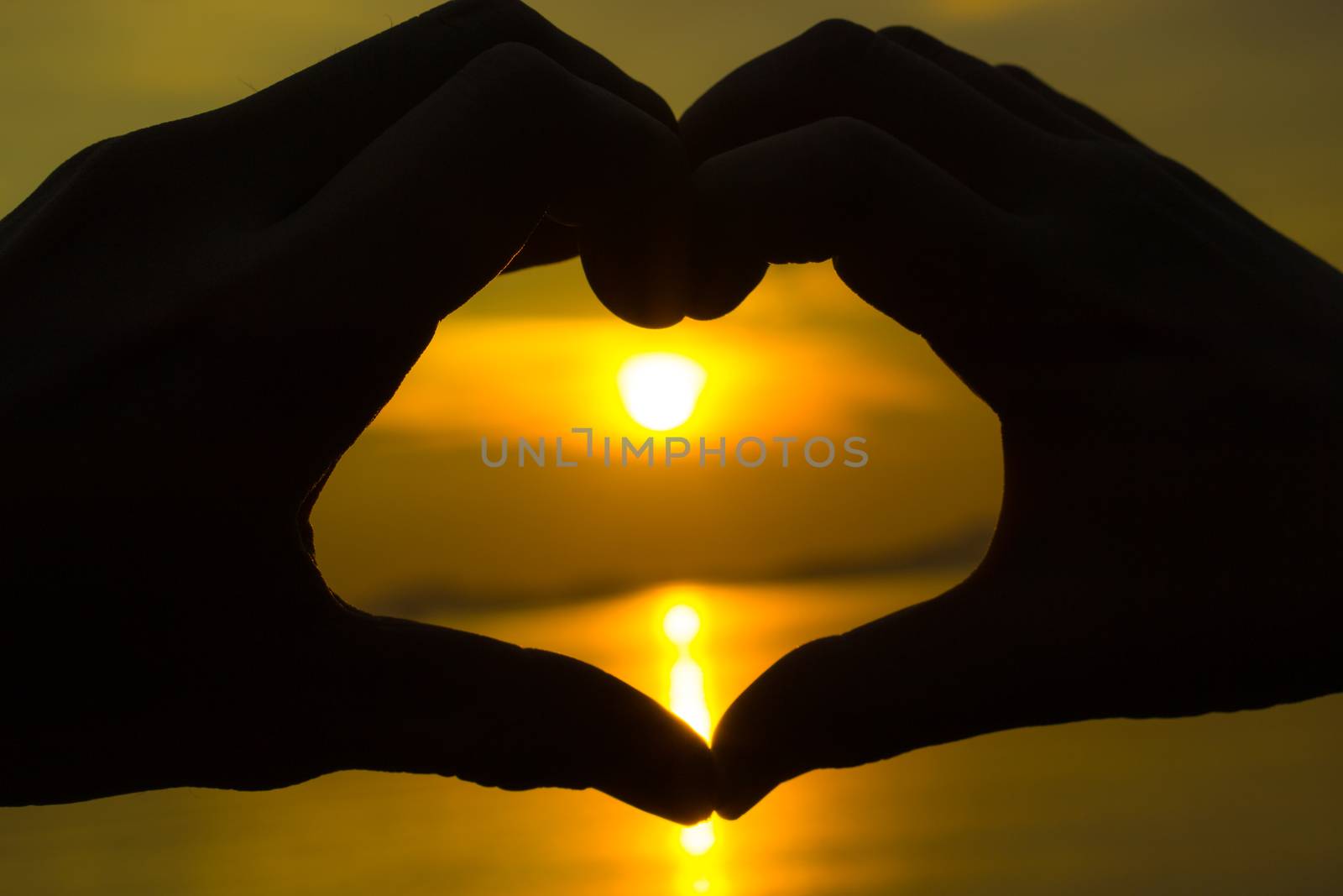 Silhouette of female hand and male hand making a heart shape in front of the sun during sunset time at the sea. Love and valentine concept.