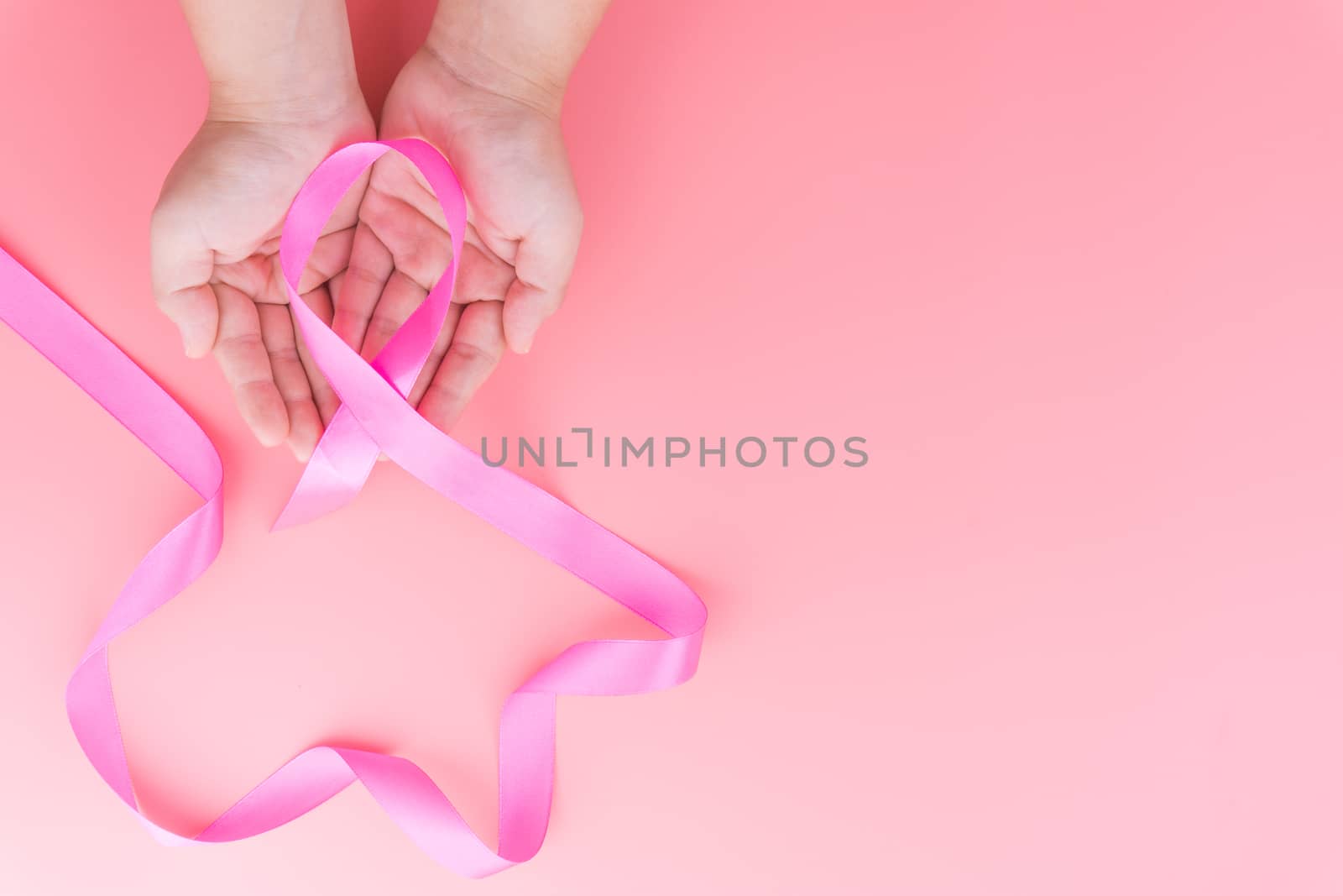 Woman hands holding pink ribbon breast cancer on pink background with copy space for text. Flat lay, top view.