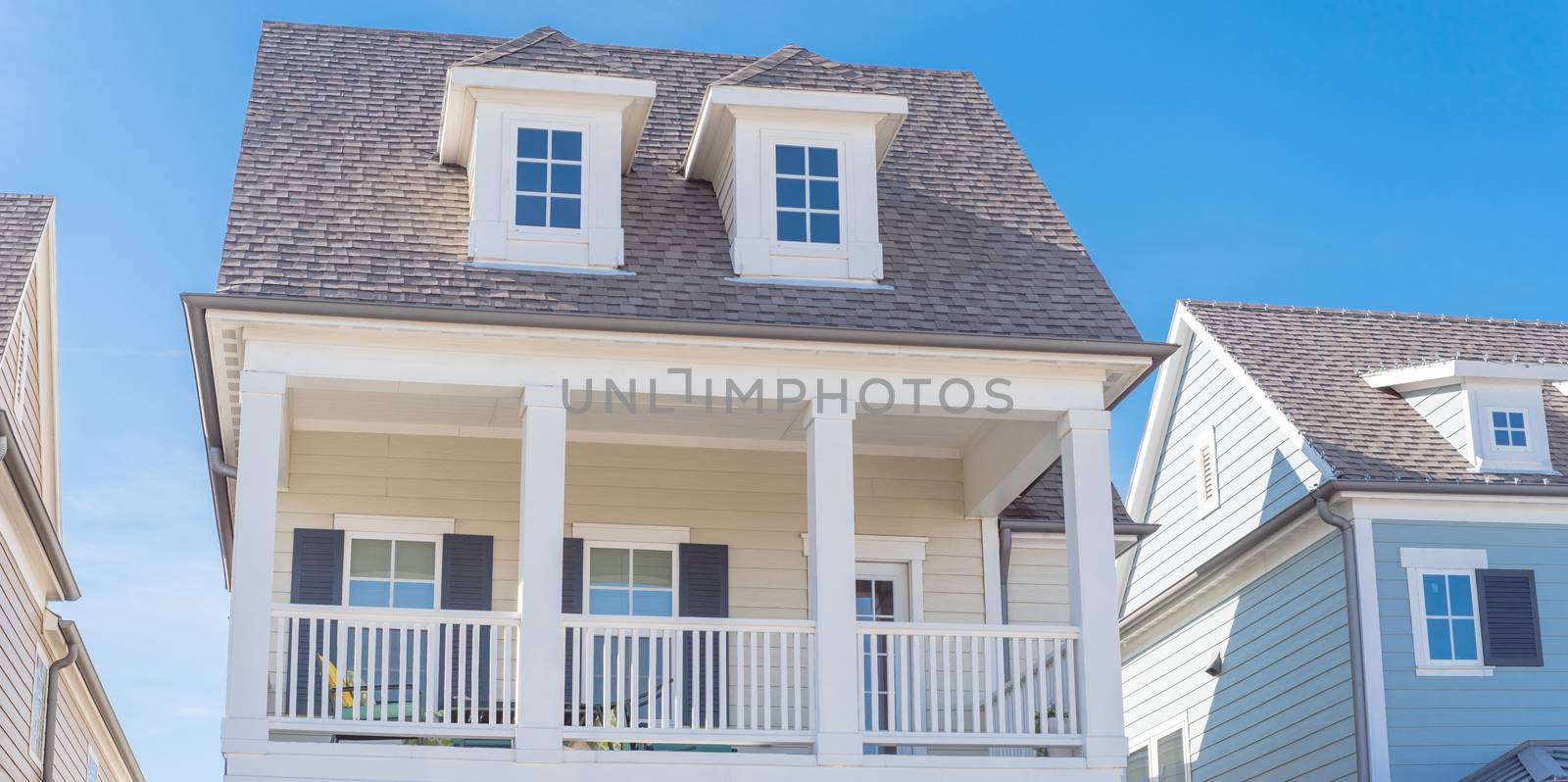 Panorama view second story porch with white painted outer edge and banister. Dormer roof windows with weathered shingle siding tiles under cloud blue sky. Country –style houses in suburbs Dallas
