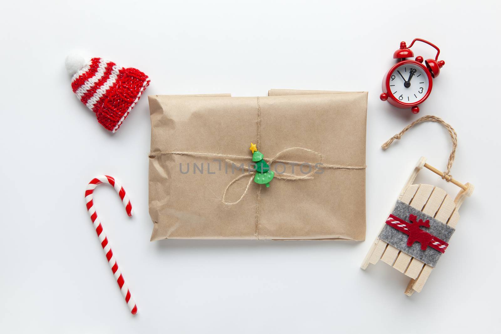 Christmas gift wrapped in brown craft paper, tied with scourge, with cane candy, small analog clock, sled, hat on white background. Minimal style. Top view. Do it yourself, celebration, eco concept.