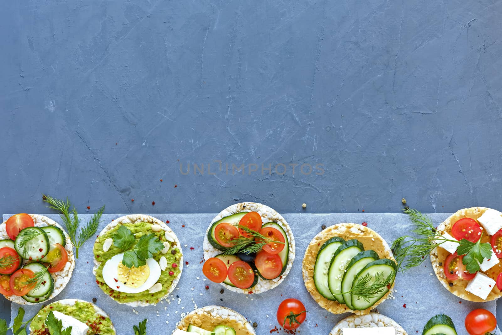 Border of savory appetizers or canapes with fresh vegetables, feta cheese and egg on rice cakes arranged on blue with copy space
