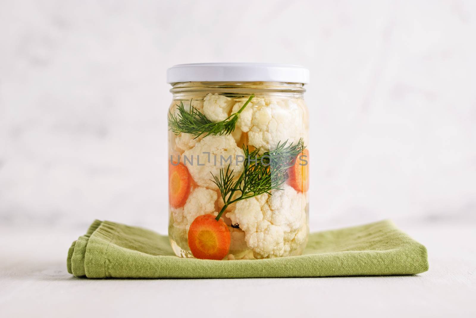 Sealed jar of fermented cauliflower with carrot and dill standing on napkin on white background