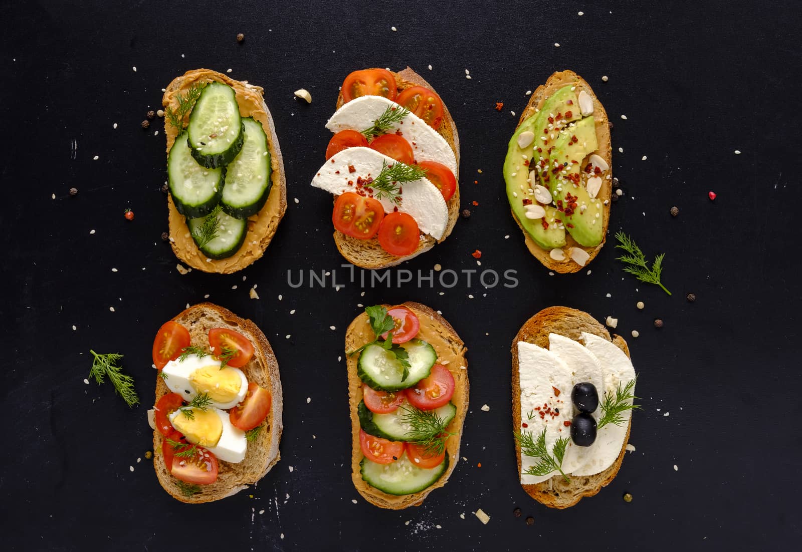 Set of fresh sandwiches snacks on dark bread with cheese, tomatoes, cucumber, boiled eggs and avocado, viewed from above on black background, decorated with greens and spices