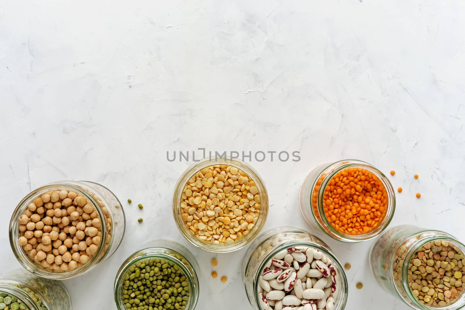 Open glass jars full of assorted dried legumes with mung beans, beans, lentils and peas over a white background in a healthy diet and nutrition concept