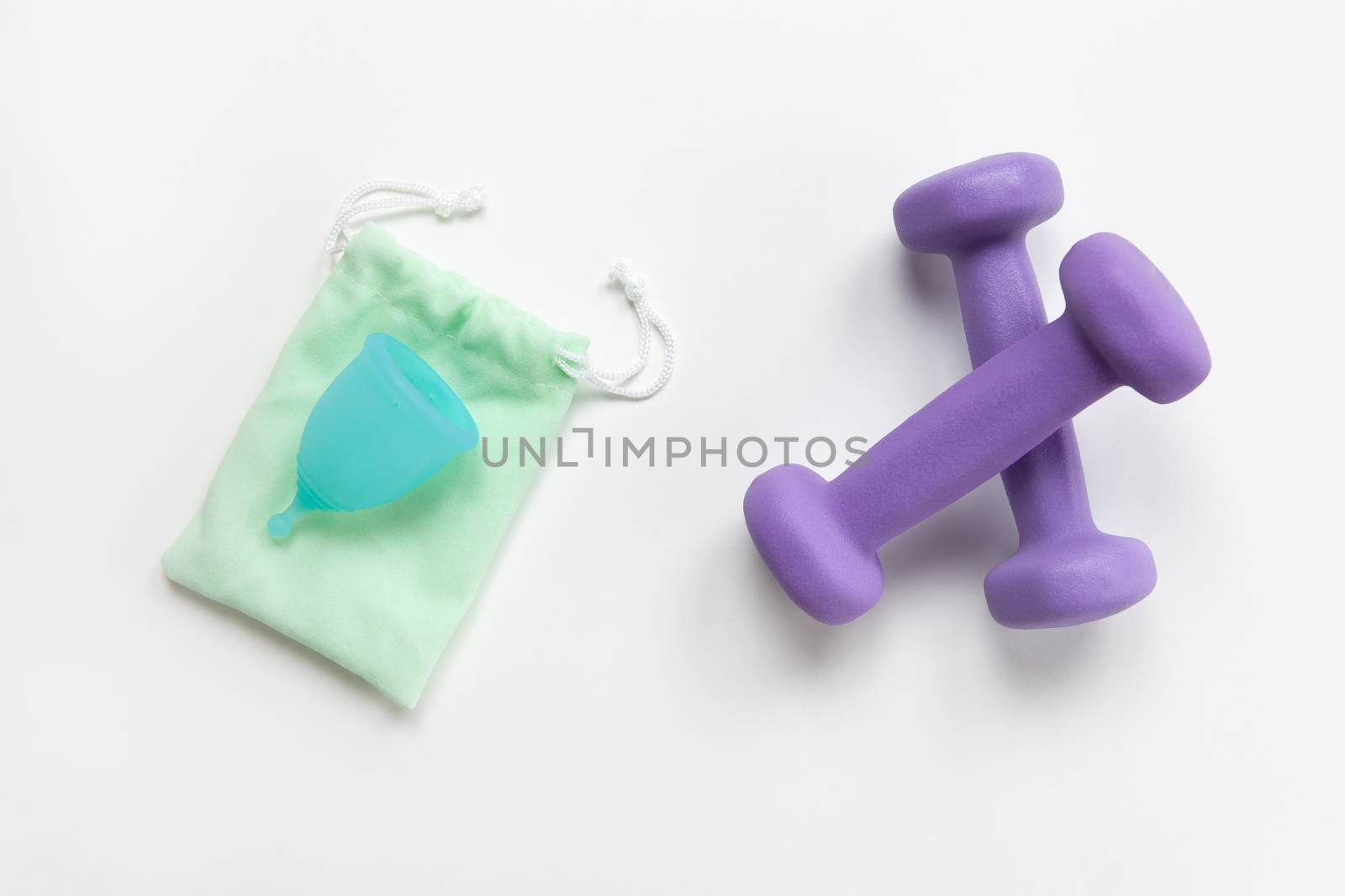 Turquoise menstrual cup on green small bag and violet dumbbells on white background. Concept zero waste, savings, minimalism, no limits. Feminine hygiene product for fitness, flat lay. Horizontal.