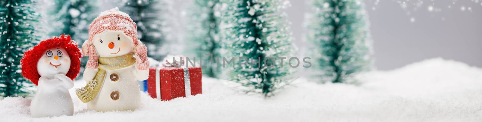 Two small cute snowmen friends with gifts in forest under falling snow