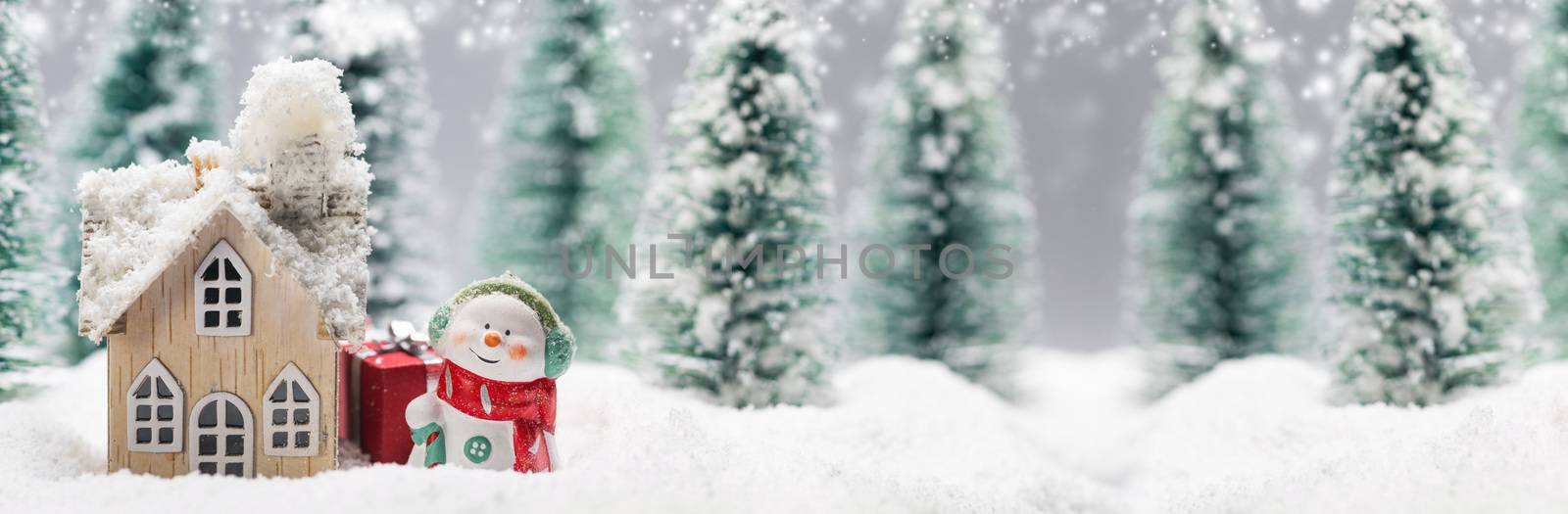 Small decorative snowman with gift near wooden house in fir forest under falling snow