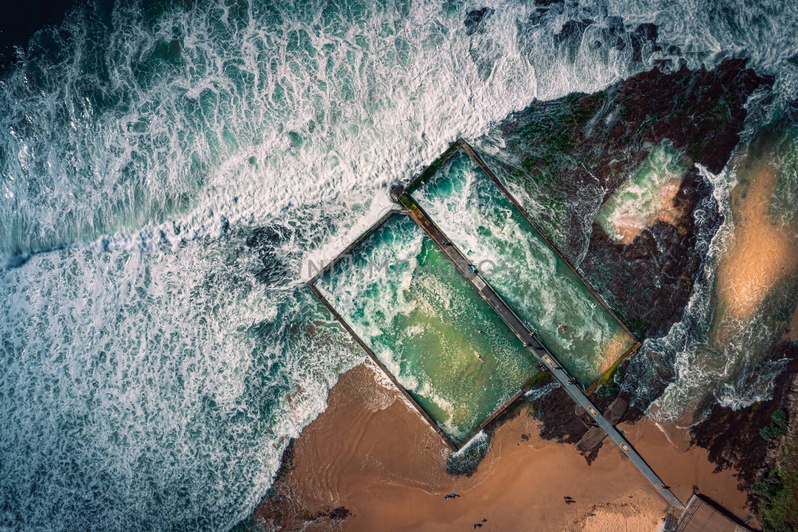 Aerial views of waves from a high tide wash over mossy rocks and the  twin ocean tidal pools at the beach.   Motion in the faster moving flows.   
Location, Austinmer, Australia