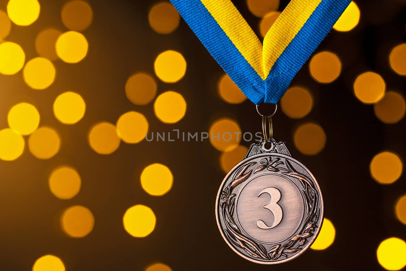 Third placed runner up bronze medallion on a ribbon hanging against a blue background with golden bokeh for a celebration of the win in a sporting competition or championship