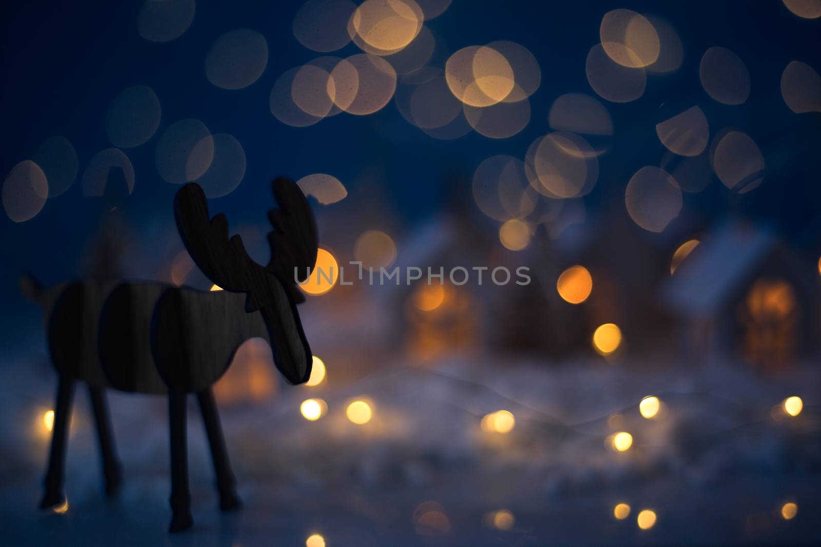 Christmas wooden reindeer over small glowing toy houses village garland over bokeh lights background with copy space for text