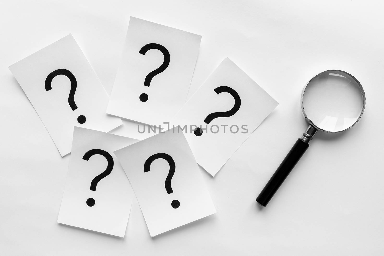 Question marks printed on white cards lying on a white background with a magnifying glass in a conceptual image