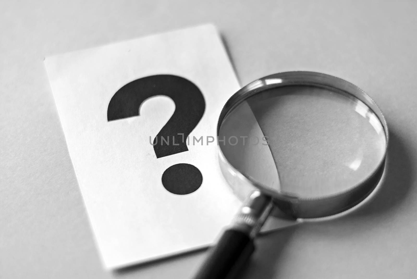 Magnifying glass on grey surface, next to big black question mark printed on white sheet of paper. Search for answers concept