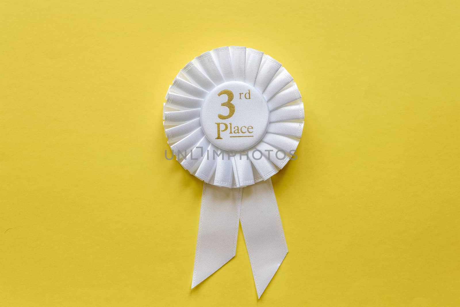 3rd place white ribbon rosette with gold text for the runner up in a competition or race on a yellow background with copy space