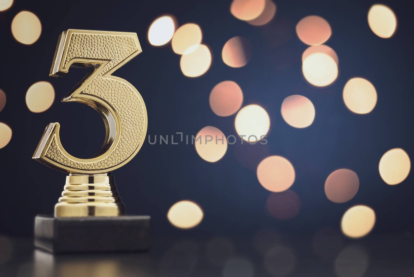 Sharp close up on single isolated gold colored plastic statue of number 3 over blue and black background and bokeh