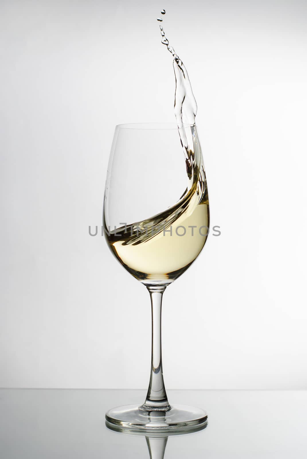 White wine splashing up the side of an elegant long stemmed wineglass in a freeze motion image on a reflective surface against a white background