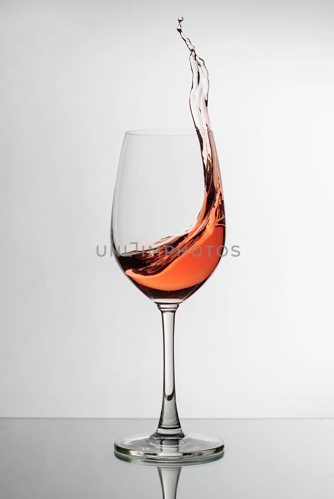 Pink wine splashing up the side of a wineglass in a freeze motion image suspended in the air over a reflective white studio background