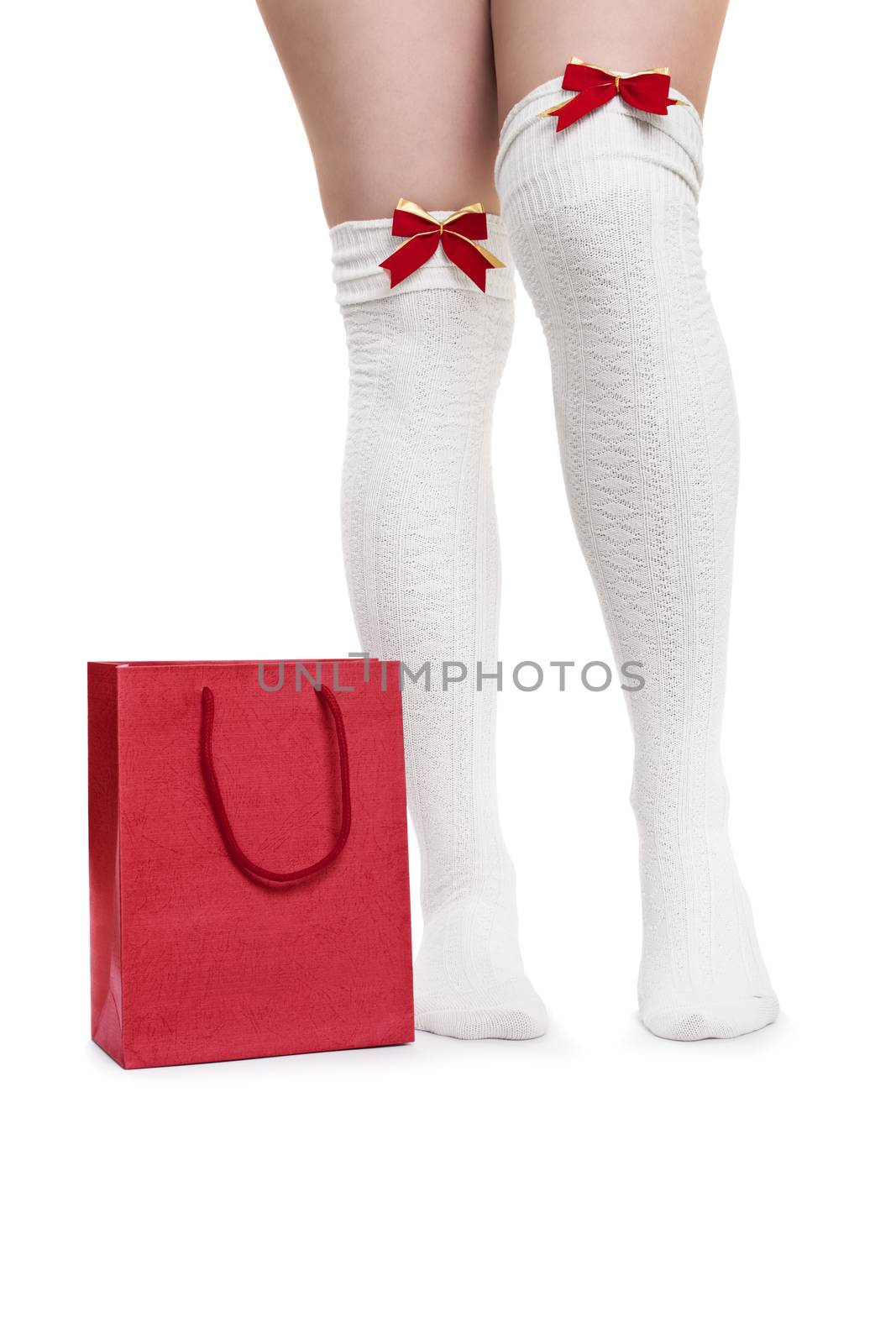 Christmas or Valentine’s Concept. Female legs in white knee high stockings with red bow ties standing next to a red gift bag or shopping bag, isolated on white background. Christmas shopping concept.