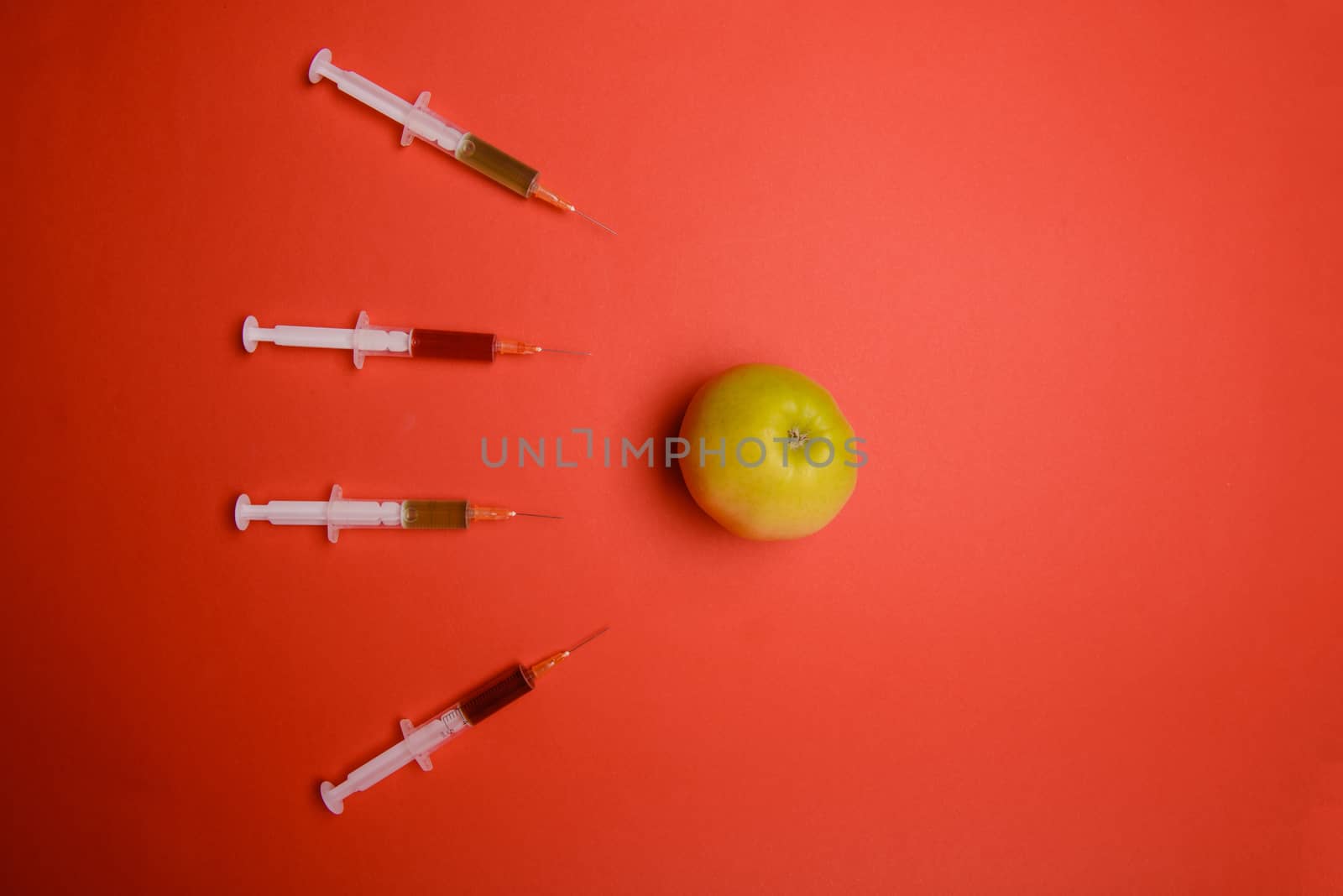 Chemical additives in food or genetically modified fruit concept. Green apple with syringes of chemicals. Isolated on red background.