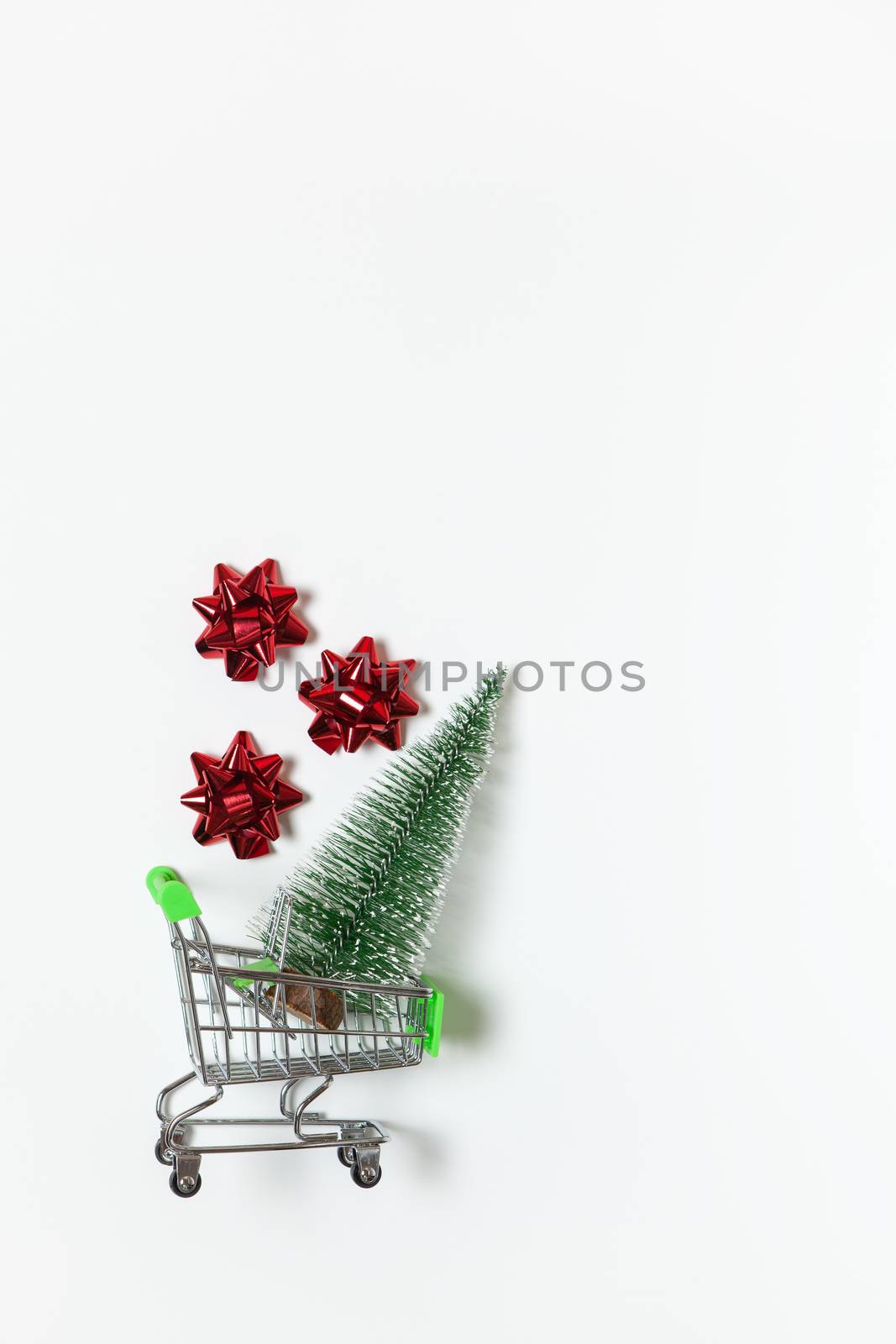 Christmas fir and red bows for gift in toy shopping cart on white background, copy space. New Year, sales, online shopping concept. Vertical, flat lay. Minimal style. Top view. For social media.