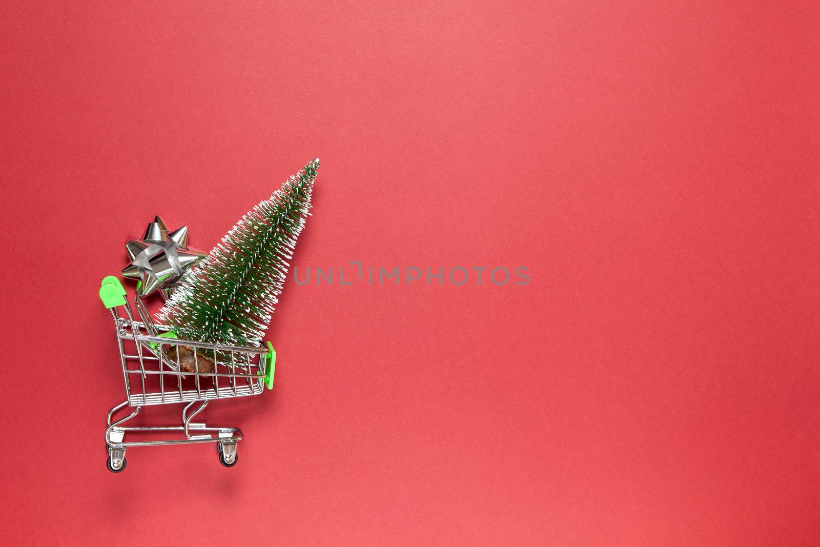 Christmas fir and silver bow for gift in toy shopping cart on burgundy background, copy space. Festive, New Year, sales, online shopping concept. Horizontal, flat lay. Minimal style. Top view.