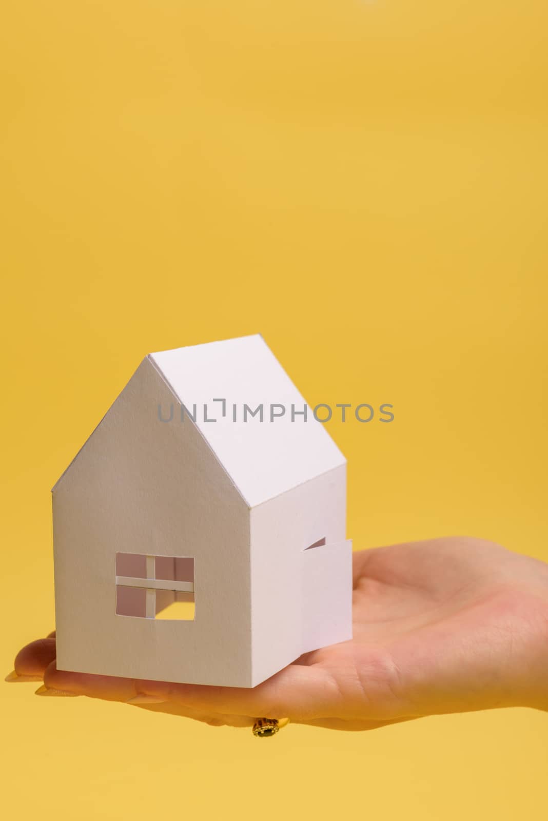 White family paper house in man hand on yellow background paper. Minimalistic style. Copy space. View from above. Vertical orientation