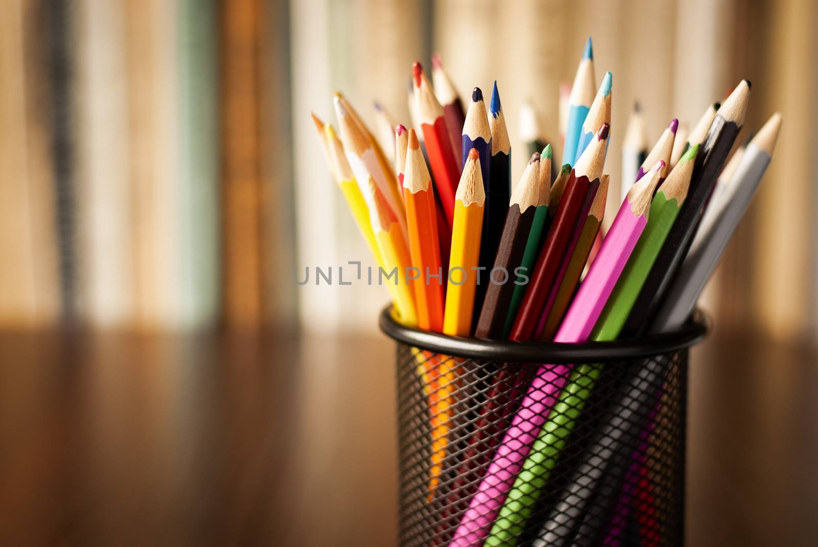 Wire desk tidy full of coloured pencils standing on a wooden table in front of a bookshelf full of books with shallow dof and copyspace