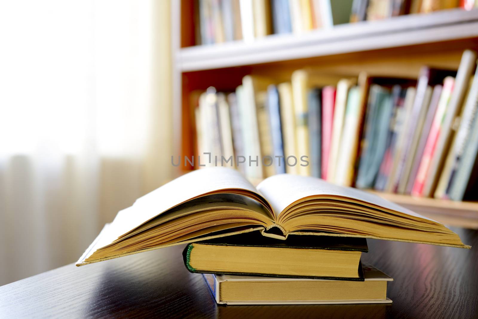 Close-up of an open book on a pile of old books, on a wooden table, with bookshelves in the background, as an invitation to study literature