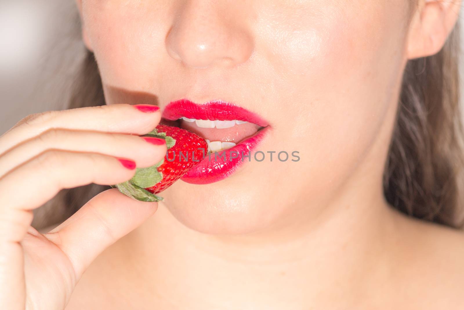 Close up of a woman with red lips and nails, biting a ripe strawberry