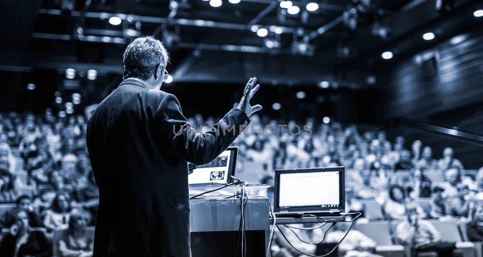 Speaker giving a talk on corporate business conference. Unrecognizable people in audience at conference hall. Business and Entrepreneurship event. Black and white, blue toned image.