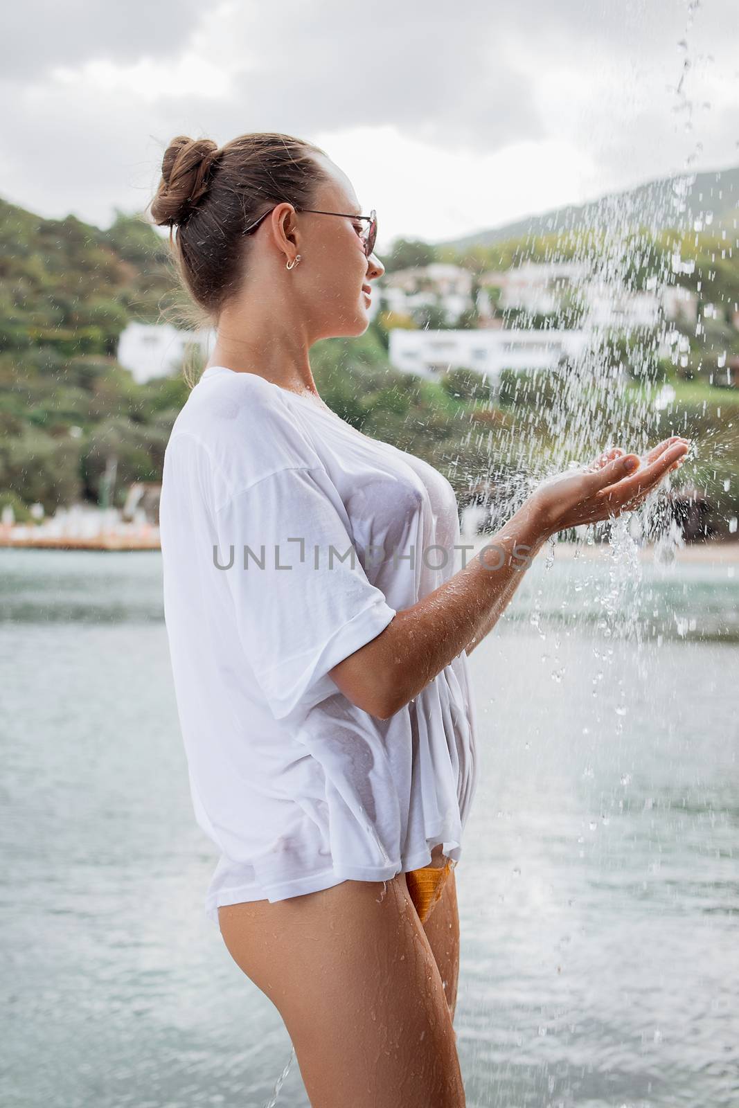 Attractive young female in wet blouse looking away while standing near splashing clean water in resort