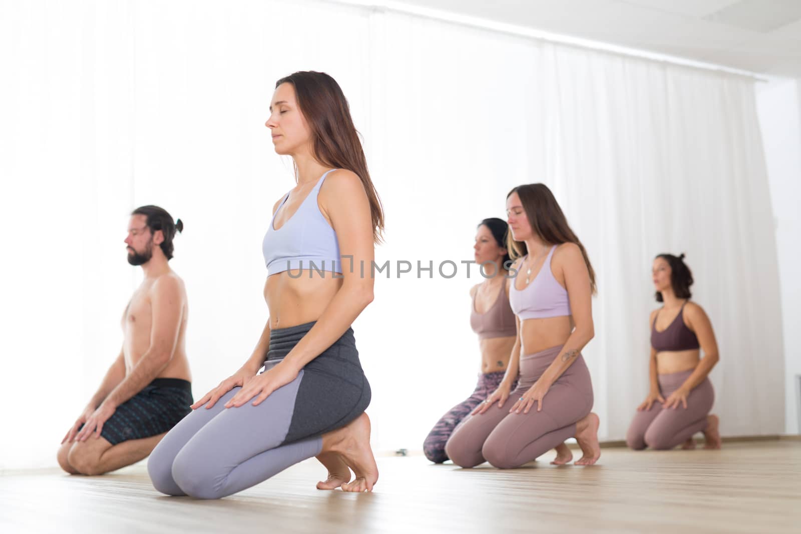 Group of young sporty attractive women in yoga studio, practicing yoga lesson with instructor, kneeling down, stretching and relaxing after workout . Healthy active lifestyle, working out in gym