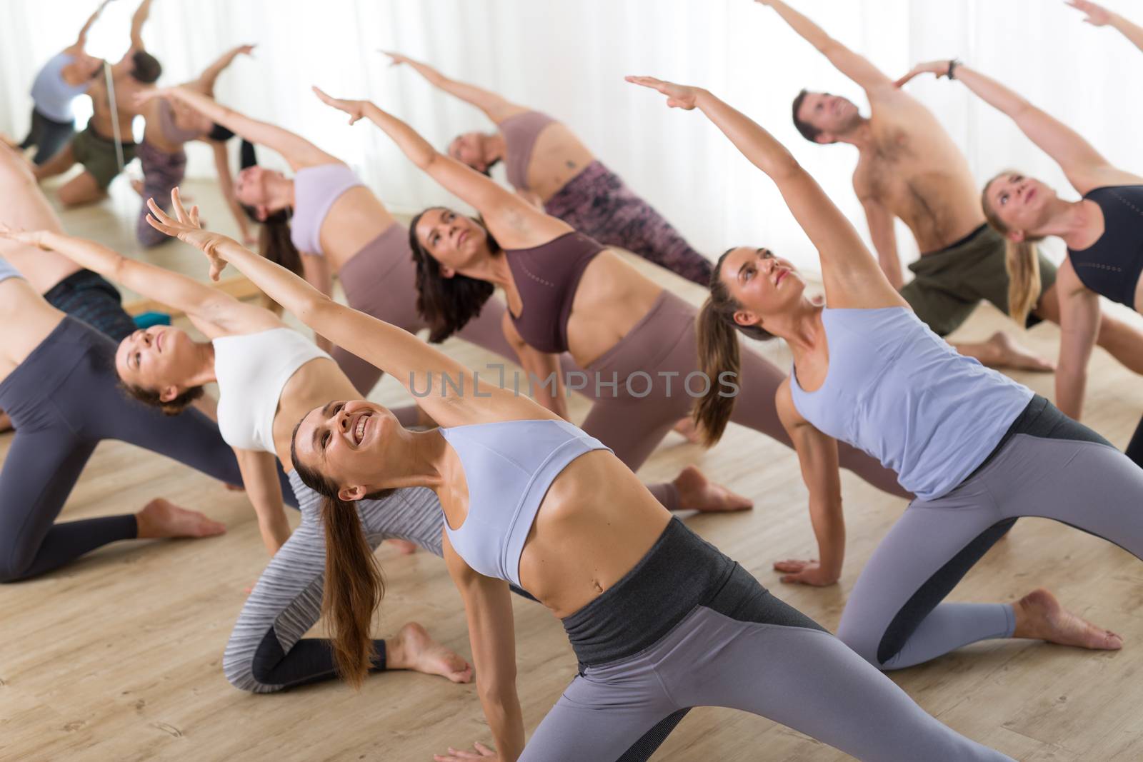 Group of young sporty attractive people in yoga studio, practicing yoga lesson with instructor, stretching on floor in Trikonosana, triangle yoga pose. Healthy active lifestyle, working out in gym. by kasto
