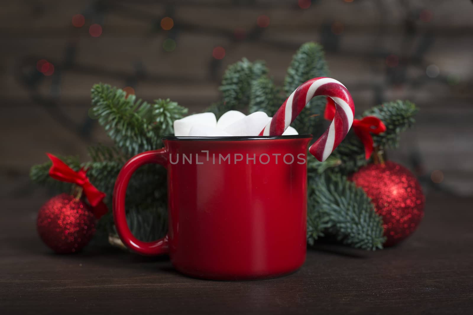 Cocoa in red mug with marshmallows candy cane fir tree branches and red baubles on dark wooden background with bokeh lights
