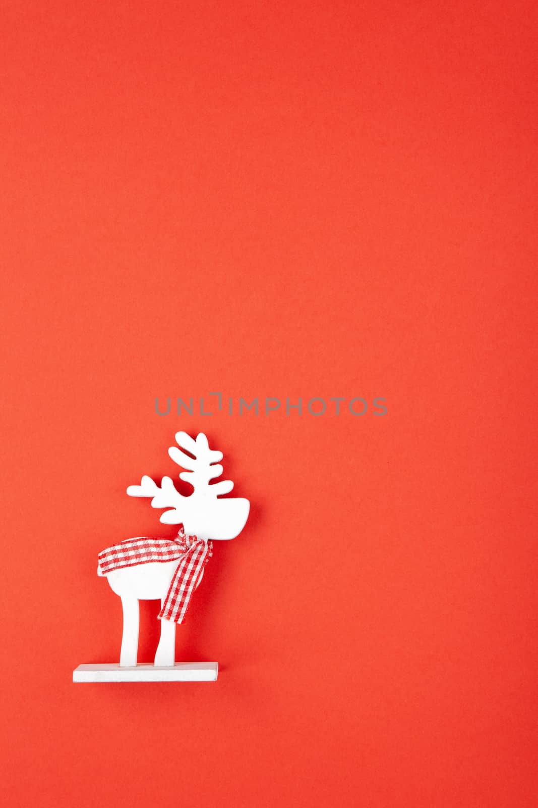 Christmas decoration, toy white deer in checkered scarf on red background, copy space. Festive, New Year concept. Vertical, flat lay. Minimal style. Top view.
