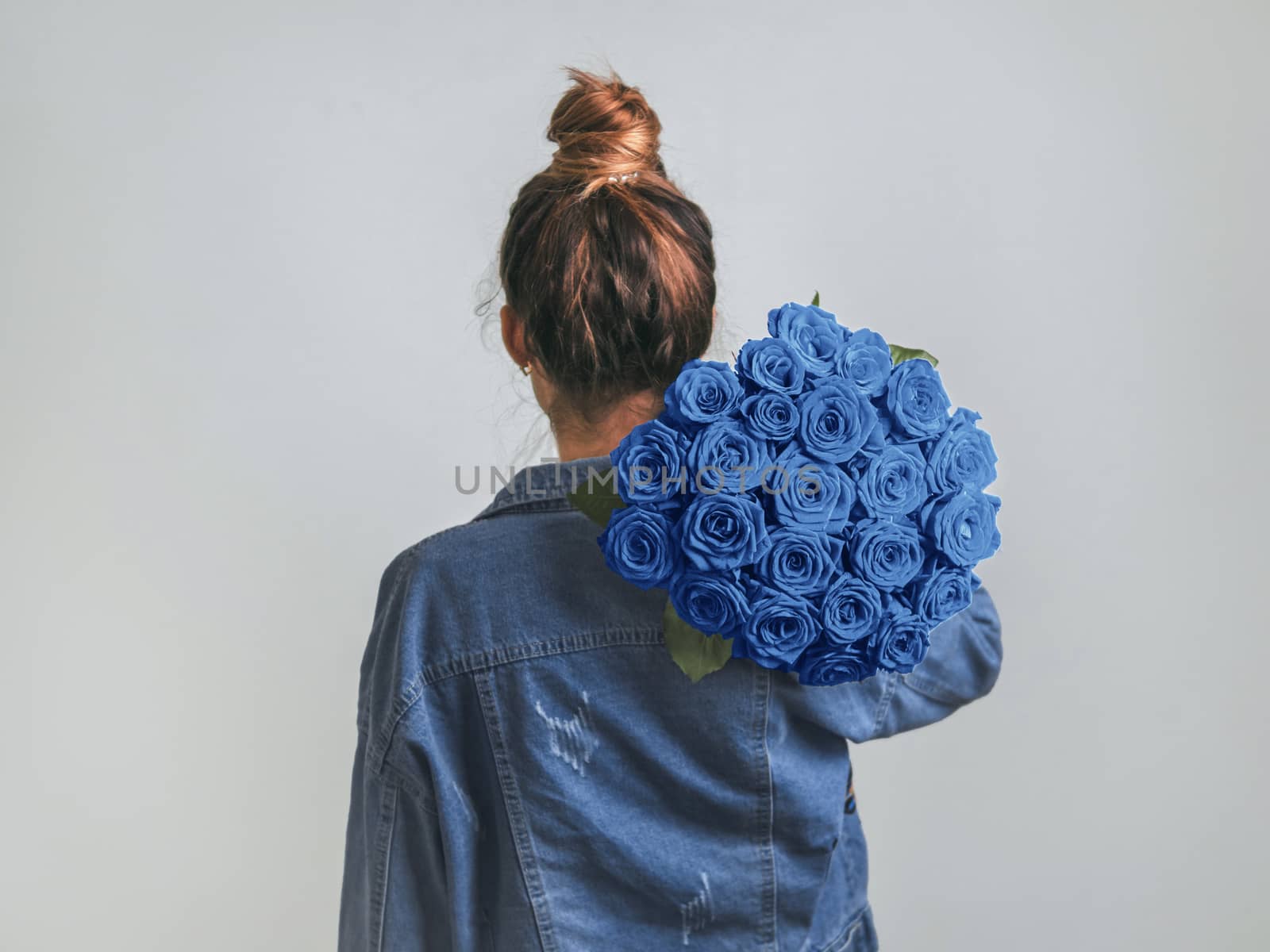 Back view of young woman in denim jacket holding bunch of Classic Blue roses on shoulder. Girl with bun updo in jeans holding flowers in Color of Year 2020 Classic Blue. Copy space.